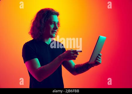 Emotive homme, étudiant avec de longs cheveux bouclés à l'aide d'un comprimé isolé sur fond dégradé jaune-magenta au néon. Concept de l'éducation, de la mode, de la jeunesse Banque D'Images
