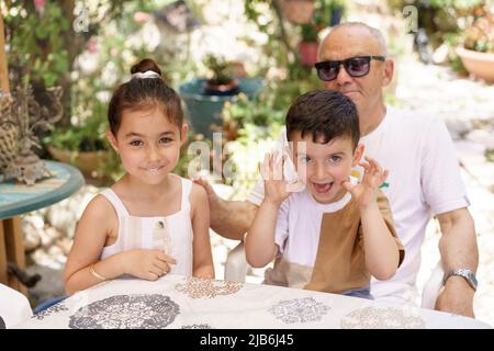 Petits-enfants heureux s'amuser avec grand-père dans l'arrière-cour, jardin d'été en arrière-plan. Petit garçon assis sur les genoux du grand-père et montrant la langue, petite fille souriant. Banque D'Images