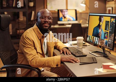 Portrait d'un graphiste africain en lunettes souriant à l'appareil photo tout en travaillant à table sur un ordinateur avec de nouveaux contenus Banque D'Images