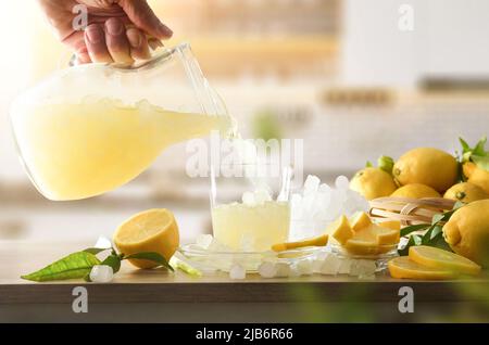 Personne versant le verre avec une boisson fraîche au citron avec de la glace du pichet sur la table de cuisine avec des citrons et de la glace pilée. Vue avant. Banque D'Images