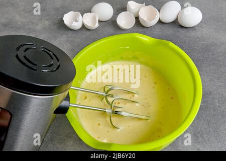 Battue les œufs avec du sucre dans un bol vert avec un batteur immergé dans un batteur et des coquilles d'œufs de poulet sur une table de cuisine en béton foncé. Création de la taille Banque D'Images