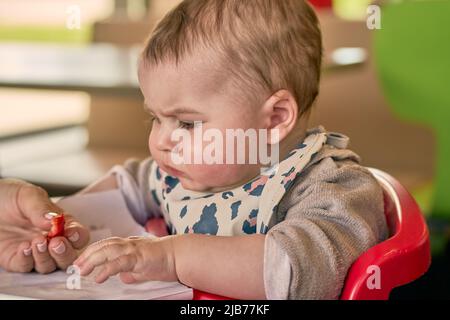 Mère donnant à bébé une tranche de fraise. Petite fille assise dans une chaise d'enfant rouge et regarde aux fruits. Elle pleure presque. Banque D'Images