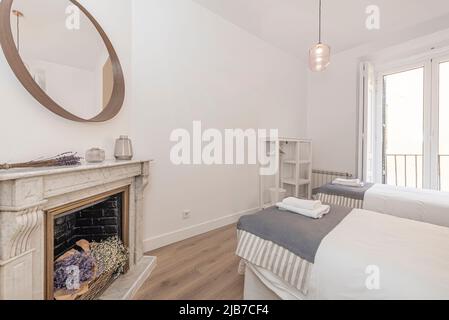 Chambre avec deux lits et cheminée en marbre blanc, miroir rond avec cadre en bois et étagère en bois blanc et balcon avec aluminium et verre Banque D'Images