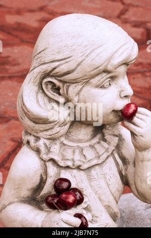 Gros plan de la statue de la petite fille montre qu'elle mange des cerises fraîches hors de sa main. La figure en ciment avec de longs cheveux regarde sur le côté. Banque D'Images