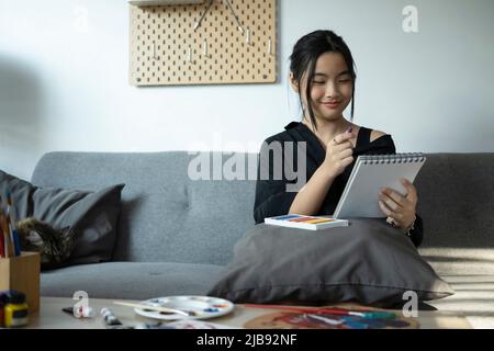 Bonne fille asiatique assise dans la salle de séjour et dessin d'image sur le carnet de croquis. Banque D'Images