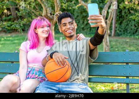 autre couple divers qui se traînait ensemble assis sur un banc de parc, faisant un selfie tenant un smartphone. adolescents interraciaux amis avec basketb Banque D'Images