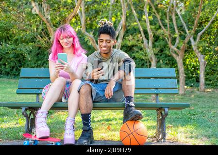 autre couple divers traînant ensemble assis dans un banc de parc tenant un smartphone en ligne sur les médias sociaux web. interracial adolescents amis Banque D'Images