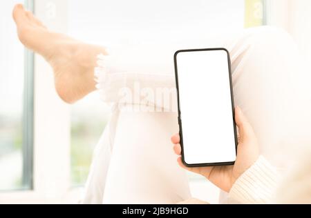 mockup image femme tenant un smartphone avec écran blanc vierge. Femme en Jean blanc est assise à pattes croisées et tient le téléphone cellulaire dans ses mains avec un blanc qui Banque D'Images