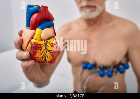 Homme senior pendant l'épreuve cardiaque avec des capteurs tenant le modèle anatomique du coeur humain à la main devant lui. Concept de santé cardiaque, tests cardiaques, cardio Banque D'Images