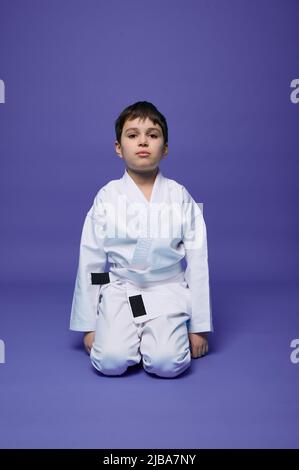 Portrait confiant d'un beau garçon d'enfant portant un kimono et assis en position aikido sur fond violet avec espace pour le texte publicitaire. Banque D'Images