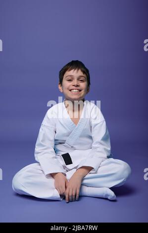 Charmant adolescent caucasien - combattant aikido - en kimono blanc sourit un sourire envoûtant, assis en position lotus tout en pratiquant les arts martiaux orientaux o Banque D'Images