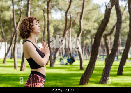 Vue latérale d'une femme gaie portant un soutien-gorge de sport noir debout sur le parc de la ville, à l'extérieur, gardez les mains dans la prière se sentent superstititititititieux religieux. Dieu. La foi, soyez Banque D'Images