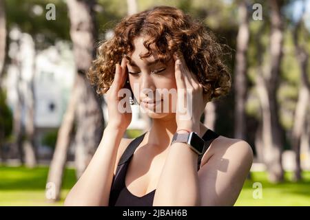 Portrait d'une jeune femme à tête rouge portant un soutien-gorge de sport debout sur le parc de la ville, en plein air tenant sa tête avec les mains essayant de se souvenir de quelque chose ou ayant h Banque D'Images