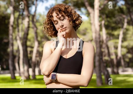 Portrait d'une jeune femme à tête rouge portant un soutien-gorge de sport debout sur le parc de la ville, extérieur touchant le menton réfléchi, pensant faire un choix important. Banque D'Images