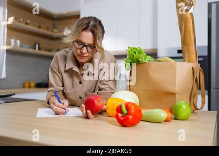 La femme au foyer mature écrit les dépenses pour la nourriture achetée. Femme dans la cuisine et un sac en papier avec épicerie Banque D'Images