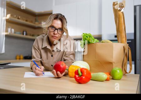 La femme d'âge moyen dans les lunettes écrit les dépenses pour la nourriture achetée Banque D'Images