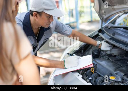 Le mécanicien automobile asiatique mâle tient un presse-papiers et examine le problème de panne de moteur de voiture avec les femmes client et d'expliquer la cause racine et d'estimer repari Banque D'Images
