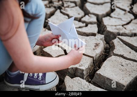 La fille abaisse le bateau en papier sur le sol sec et fissuré. Crise de l'eau et changement climatique concept. Réchauffement de la planète Banque D'Images