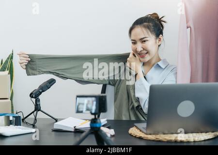 De jeunes entrepreneurs asiatiques ramassant leurs vêtements pour les montrer à leurs clients par le biais de ventes en ligne sur un téléphone en direct. Banque D'Images