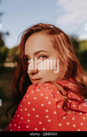 REDHEAD femme regardant la caméra au-dessus de son épaule. Femme avec de longs cheveux bouclés vêtus de robe rouge avec des points blancs Banque D'Images