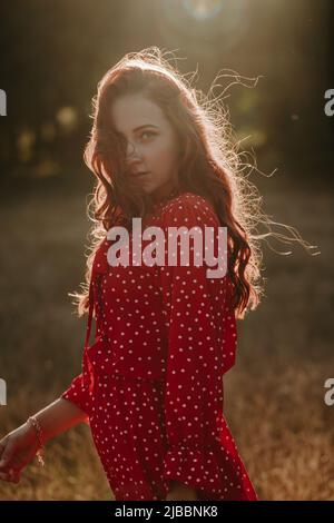 Portrait moyen de la jeune femme à tête rouge en robe rouge avec des points blancs sur fond de contre-jour de coucher de soleil. Modèle féminin regardant l'appareil photo Banque D'Images