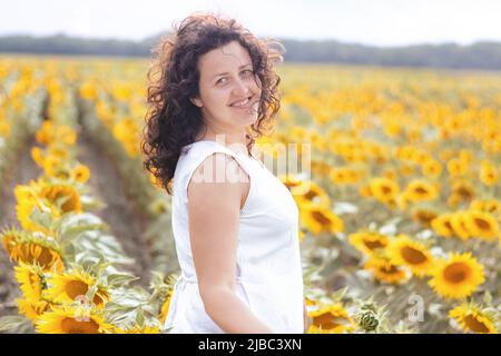 Mise au point douce sur le portrait d'une fille souriante dans le champ de tournesol. Femme aux cheveux bouclés dans la robe blanche marche. Le tournesol est l'agriculture ukrainienne. Or jaune Banque D'Images