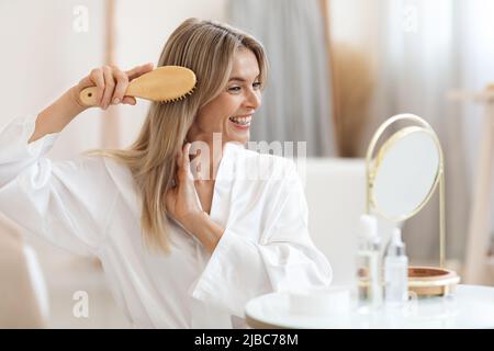 Souriante belle femme blonde peignant les cheveux à la maison Banque D'Images