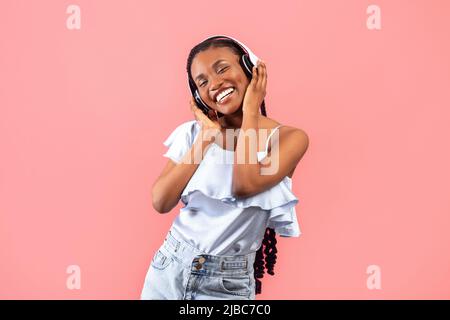 Bonne jeune femme noire dans un casque sans fil écoutant de la musique populaire sur fond rose studio Banque D'Images