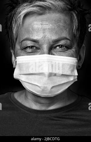 Photo en noir et blanc. Joyeux yeux sourire gris court cheveux médecin femme portant un masque médical regardant l'appareil photo portant un chemisier vert isolé sur fond jaune. Banque D'Images