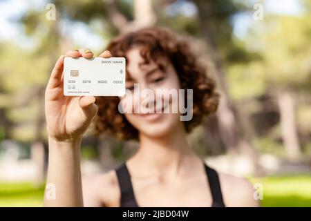 Jeune femme souriante confiante portant des vêtements de sport sur le parc de la ville, à l'extérieur montrant des cartes de crédit ou de débit en plastique, effectuant des paiements en ligne. Sélectif Banque D'Images