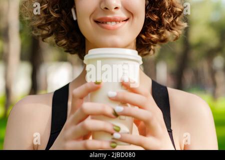 Jeune fille à tête rouge souriante heureuse portant un soutien-gorge de sport debout sur le parc de la ville, à l'extérieur tenant une tasse à café à emporter devant la poitrine et les yeux fermés. COFF Banque D'Images