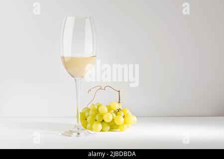 Vin blanc dans un gobelet en verre et un bouquet de raisins sur une table blanche. Arrière-plan clair. Banque D'Images