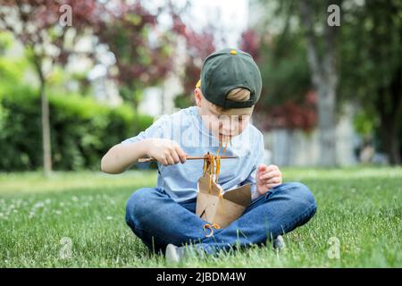 Enfant mangeant remuer les nouilles udon frites en position assise sur l'herbe. Sale tache de sauce sur son t-shirt. Des nouilles sont tombées sur ses vêtements. À l'extérieur Banque D'Images