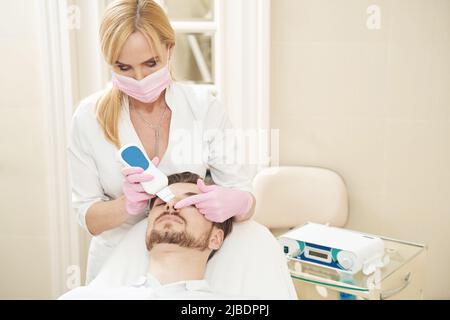 Femme ouvrier de beauté traitant le nez de l'homme avec l'épurateur de peau ultrasonique Banque D'Images