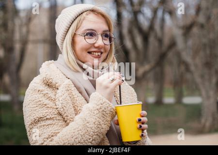 Joli blond décontracté dans le parc, souriant et buvant du café à emporter. Jeune femme moderne tenant une tasse jaune dans sa main. Banque D'Images