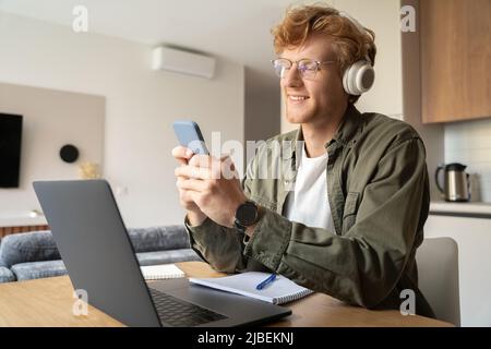 Jeune Ginger homme utilisant un smartphone tout en travaillant en ligne sur un ordinateur portable au bureau à domicile Banque D'Images