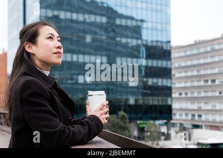 Vue latérale d'une femme d'affaires chinoise pensive dans un vêtement d'extérieur chic et décontracté avec une tasse de boisson chaude écologique. Elle regarde loin et pense en se tenant sur le pont avant de travailler le matin dans le centre-ville Banque D'Images