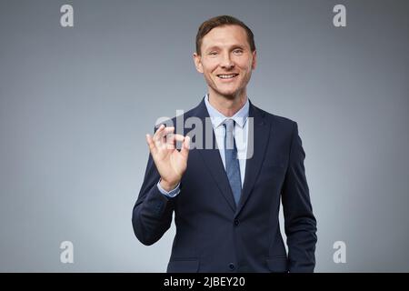 Portrait à la taille de l'homme d'affaires caucasien souriant montrant le signe OK tout en se tenant contre fond gris dans le studio, espace de copie Banque D'Images