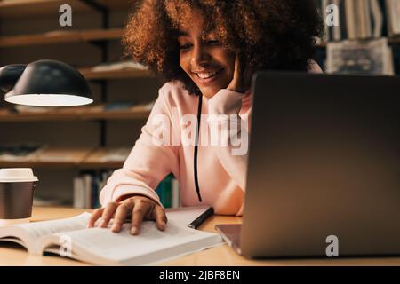 Étudiant souriant étudiant tard dans la nuit. Jeune femme utilisant un livre et un ordinateur portable au bureau de la bibliothèque. Banque D'Images