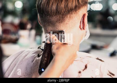 Boutique de photos Lifestyle. Jeune homme, le client porte un masque, vue arrière latérale arrière. Tondeuse électrique à cheveux pour coiffeur, homme, main, travail, peigne Banque D'Images