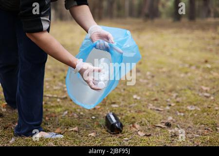 Une femme tient les mains dans des gants pour recueillir et mettre la bouteille de plastique usagée dans un sac poubelle bleu. Un volontaire nettoie le parc par une journée ensoleillée. Effacement, p Banque D'Images