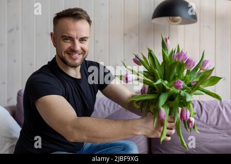 Jeune homme beau est assis à la maison sur un canapé avec bouquet de tulipes et prépare la surprise pour sa petite amie. Préparation de la date. Attention et soin Banque D'Images
