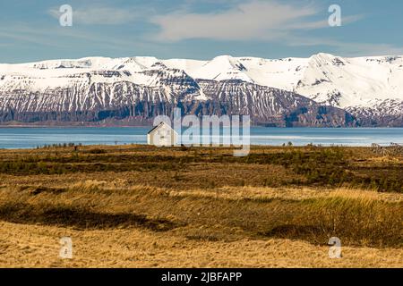 Une maison en bois blanche se dresse sur une langue de terre près de Húsavík, dans le nord de l'Islande, face à des montagnes enneigées au-dessus de la baie de Skjálfandi. Des séchoirs en bois appartenant à l'exploitation aquacole Fiskeldið Haukamýri sont visibles à droite, utilisés pour sécher le poisson. Banque D'Images
