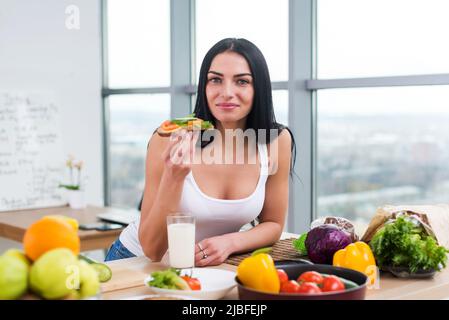 Gros plan de femme souriante mangeant un sandwich végétarien avec des légumes pour le petit déjeuner le matin, regardant l'appareil photo Banque D'Images