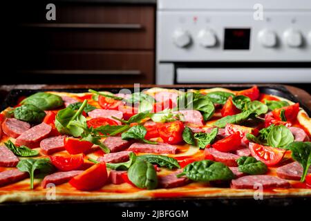 Faire cuire de la pizza avec des saucisses, des tomates, des feuilles vertes en gros plan sur une plaque de cuisson dans la cuisine. Banque D'Images