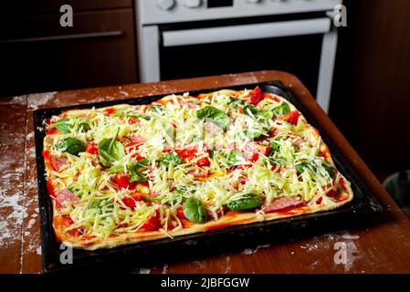 Faire cuire de la pizza avec des saucisses, des tomates, des feuilles vertes en gros plan sur une plaque de cuisson dans la cuisine. Banque D'Images