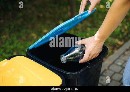 Gros plan portrait femme main throwig bouteille de verre vide dans le bac de recyclage. Différentes couleurs de bacs en plastique dans le parc. Couvercle bleu Banque D'Images