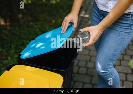 Gros plan portrait femme main throwig bouteille de verre vide dans le bac de recyclage. Différentes couleurs de bacs en plastique dans le parc. Couvercle bleu Banque D'Images