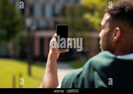 Homme afro-américain tenant un smartphone avec un écran vierge, prenant des photos ou ayant des appels vidéo à l'extérieur, mockup Banque D'Images