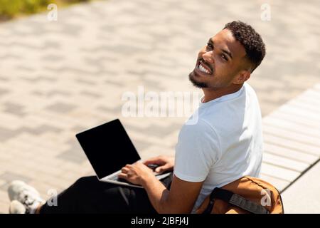 Au-dessus de la vue d'un homme afro-américain heureux en utilisant un ordinateur personnel assis à l'extérieur dans la ville, mockup Banque D'Images